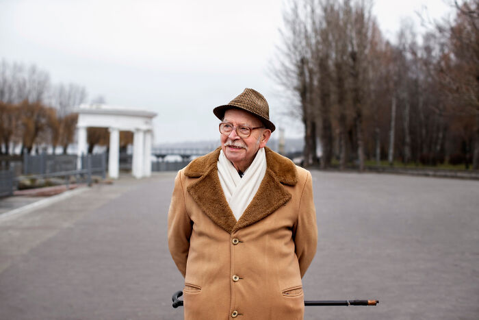 Elderly man in coat and hat, standing outdoors with cane, illustrating aging realities.