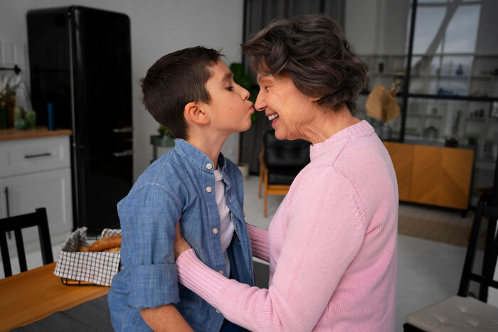 Child jokingly whispers a bizarre house rule to a senior woman, both smiling in a cozy kitchen.