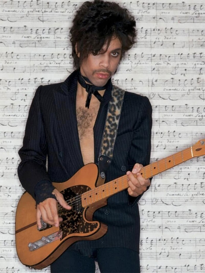 Musician with guitar in front of musical notes, wearing a dark jacket and leopard print strap.