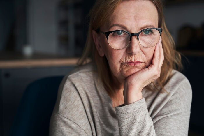 Elderly woman wearing glasses, resting her chin on her hand, reflecting on aging realities.