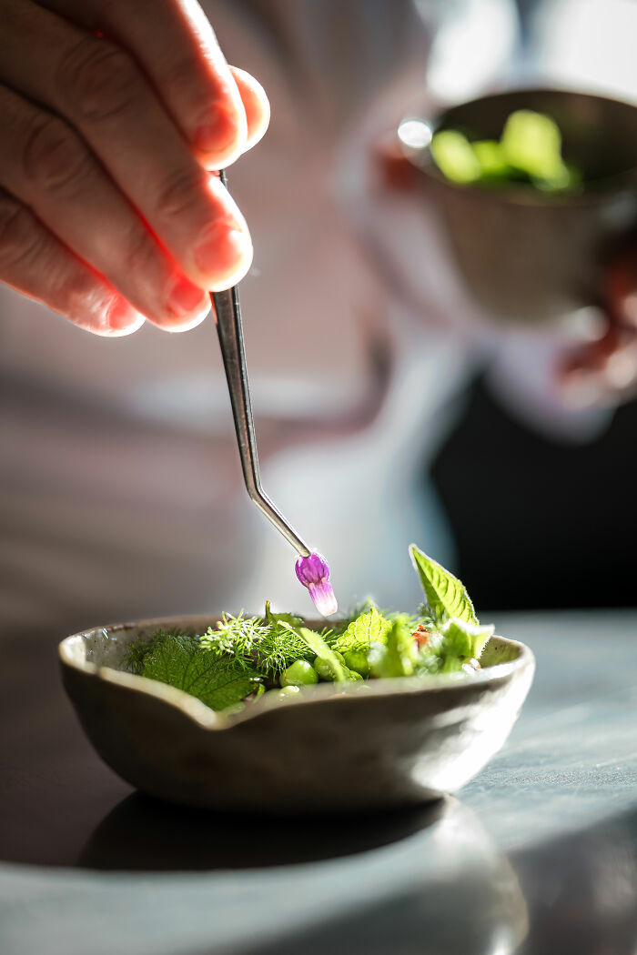 Chef garnishing dish with tweezers, featuring fresh greens and a small purple flower, from 2025 World Food Photography Awards.