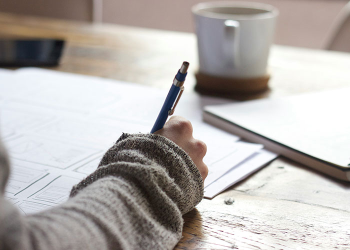 Person writing notes with a blue pen on paper, a mug on the desk, symbolizing teacher activities.