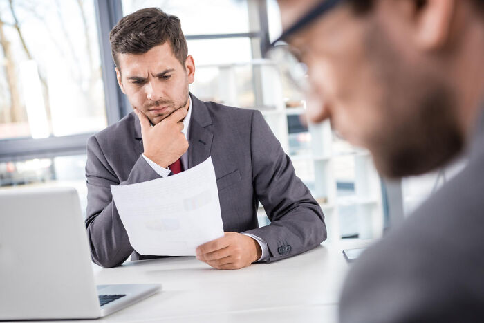 Man in a suit reviewing papers thoughtfully during a job interview with focus on ridiculous questions asked.