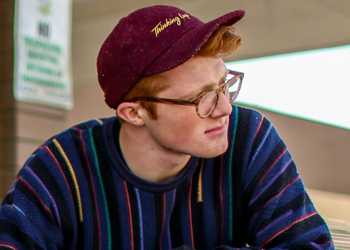 A person in glasses and a striped sweater, wearing a maroon cap, looking thoughtful.