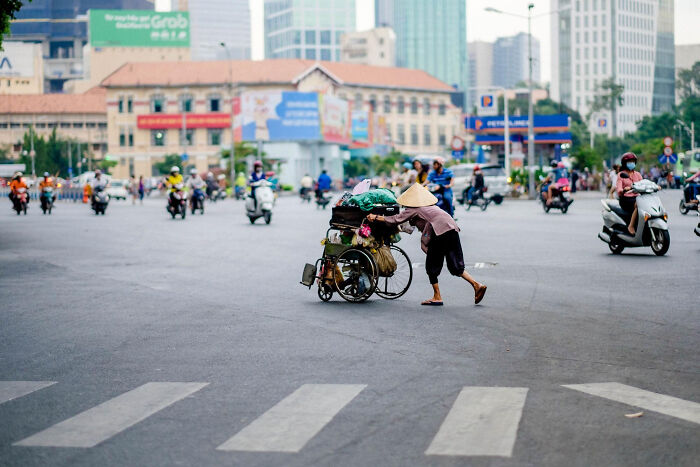 Person wearing a conical hat pushing a loaded wheelchair across a busy street in a super normal thing country setting.