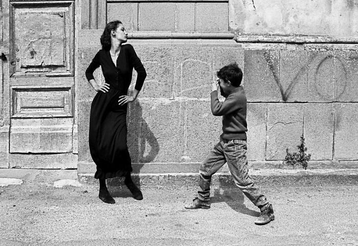 Black and white street photo of a woman posing confidently while a child playfully faces her on a city sidewalk.