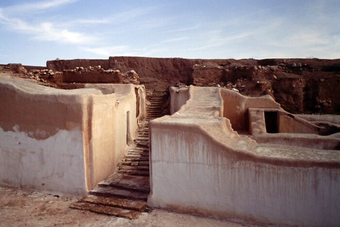 Ancient man-made structures with eroded walls and stone steps under a partly cloudy sky in a desert setting.