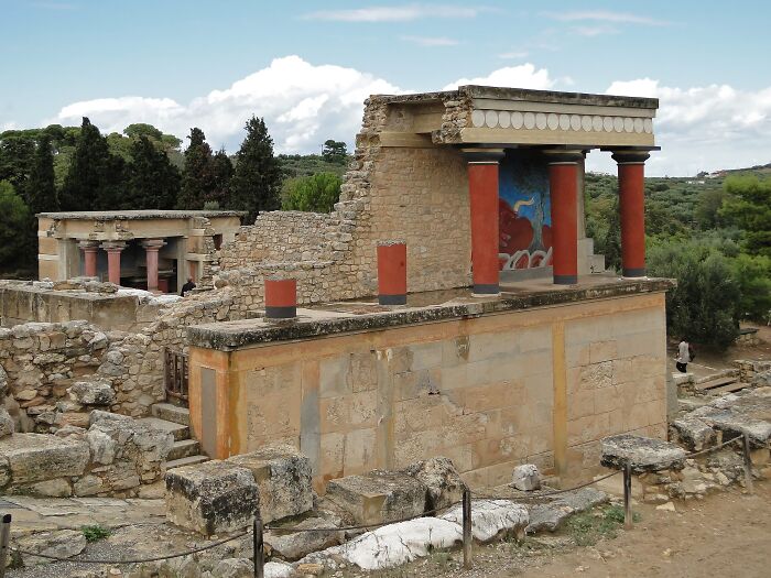 Ruins of an ancient architectural wonder with red columns and stone walls under a cloudy sky, surrounded by greenery.