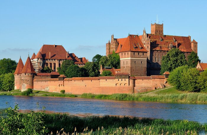 Historic architectural wonders with red brick towers and walls by a river, surrounded by greenery.