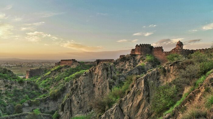 Ancient architectural wonder on a rugged landscape at sunset, surrounded by cliffs and greenery.