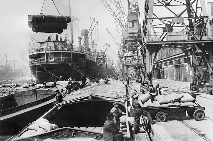 Workers unloading cargo from ships at a busy WWII dockyard, surrounded by cranes and large vessels.