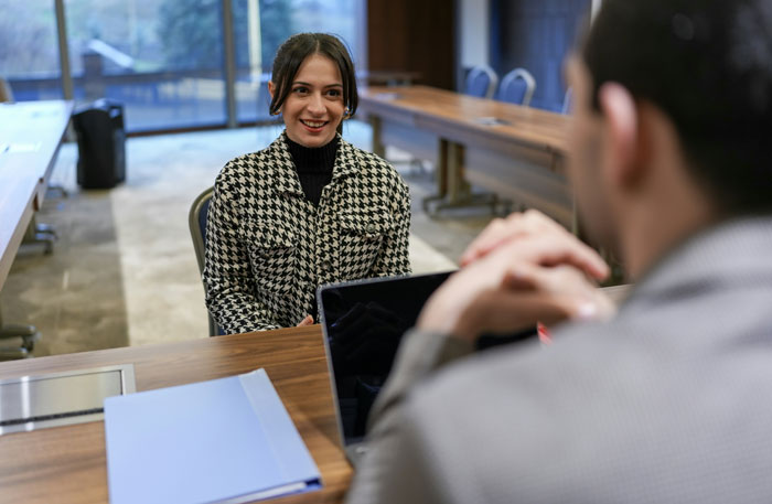 Job hunting interview with a woman in houndstooth jacket, smiling across a table with a laptop and documents.