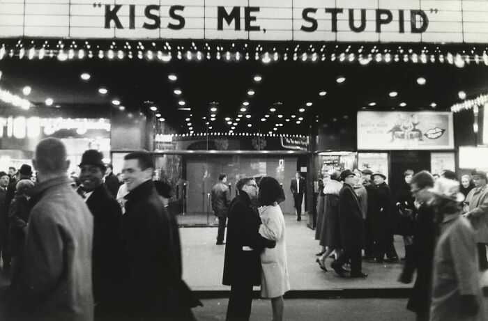 Black and white street photo showing a couple kissing under a lighted marquee with pedestrians walking by.