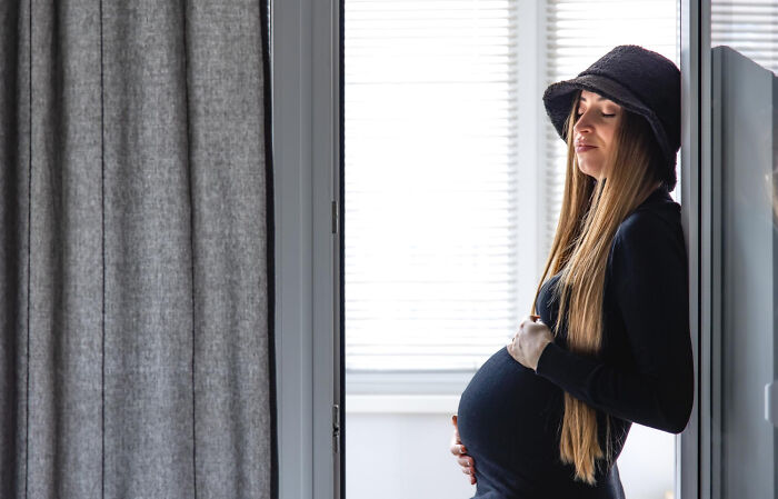 Pregnant woman in a black hat and sweater, standing by a window, eyes closed, in a calm moment. High school memories.