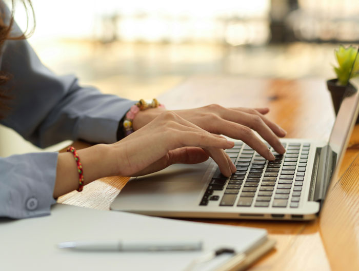 Person typing on a laptop keyboard at a wooden desk, focusing on submitting SEO deliverables in a creative way.