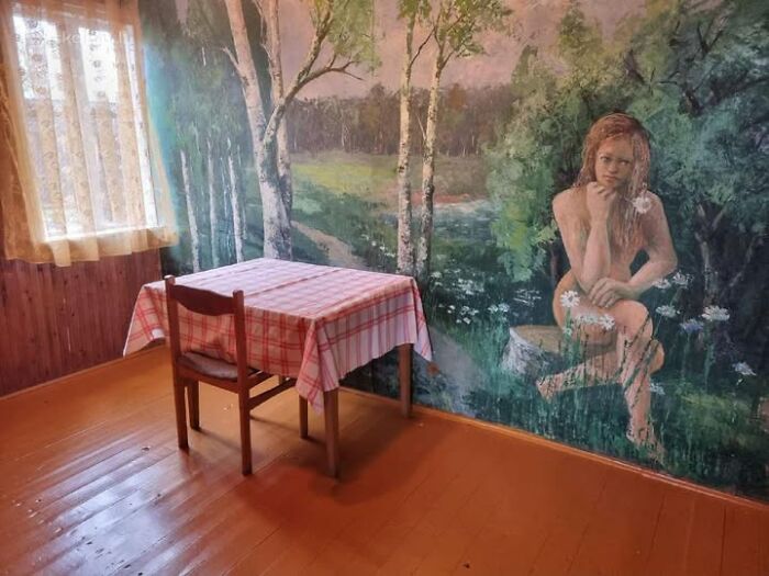 Simple room with wooden floor, small table with checkered cloth, and large mural of a woman in nature, wild real estate property.