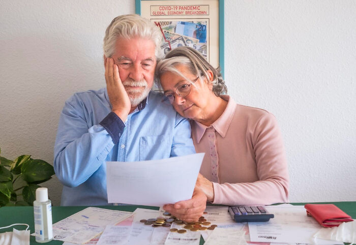 Elderly couple reviewing documents with concern, dealing with financial issues, COVID-19 poster in background, therapy support.