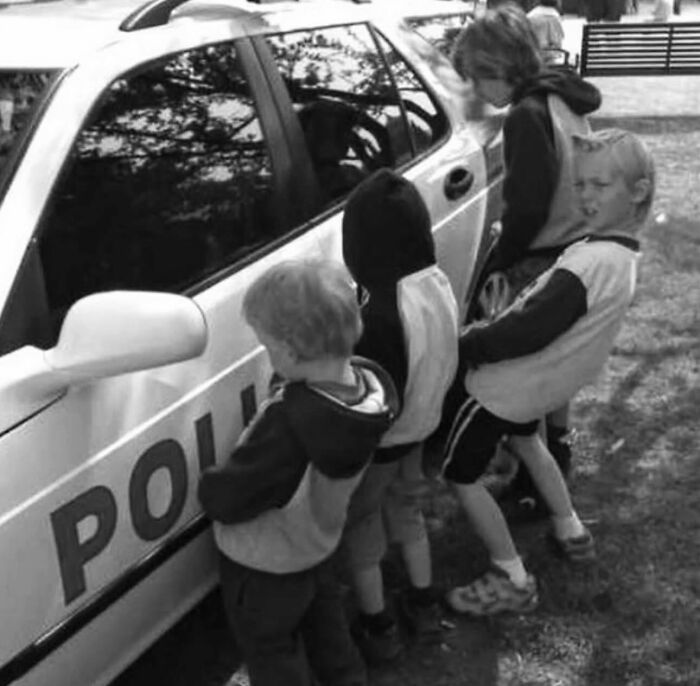 Four children in black and white street photo standing next to a police car, captured in candid outdoor moment.