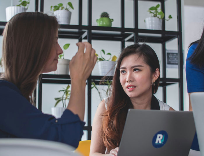 Two women in a modern office setting discussing SEO deliverables with laptop and plants in the background.
