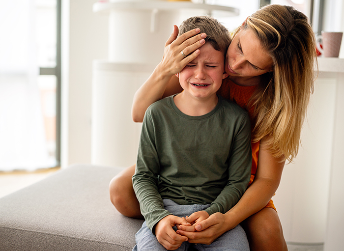 A concerned mom comforts her crying child, sitting on a couch in a bright room.
