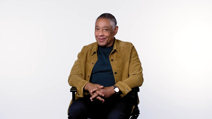 Celebrity in a tan jacket sits smiling in a chair against a plain background.