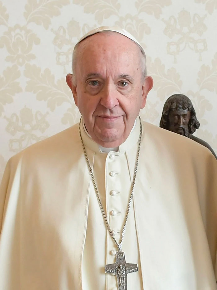 Pope Francis standing in traditional attire, with a solemn expression, wearing a cross necklace.