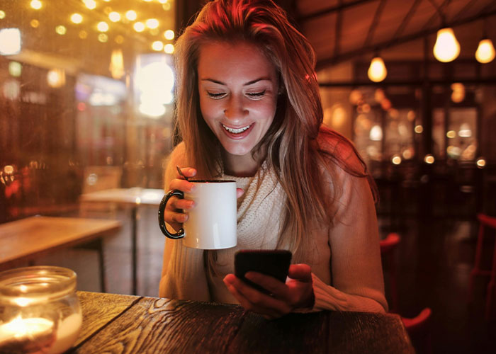 Woman in cozy café, smiling at her phone while holding a coffee mug, under warm lights.