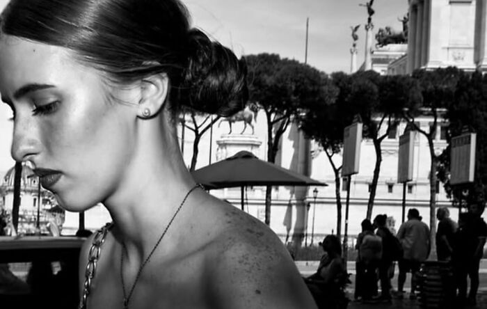 Black and white street photo of a woman with hair in a bun, captured on a busy urban street scene with trees and people in background