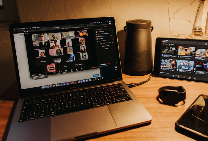 Laptop showing a virtual meeting with multiple participants, next to a tablet, speaker, smartwatch, and phone on a wooden desk.