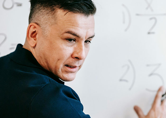 A teacher points at math equations on a whiteboard, emphasizing a lesson in a classroom setting.