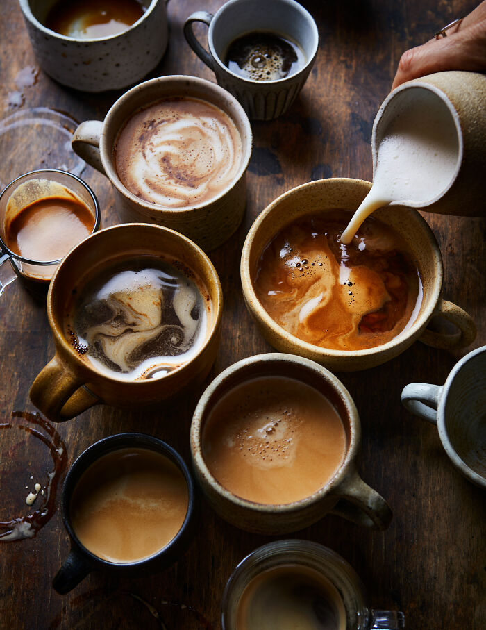 Assorted coffee mugs with varied foam designs on a wooden surface, showcasing culinary artistry from World Food Photography Awards.