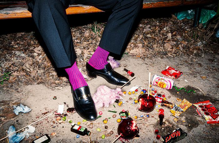 Man in purple socks, black shoes among scattered candy and alley debris, representing World Food Photography Awards entry.