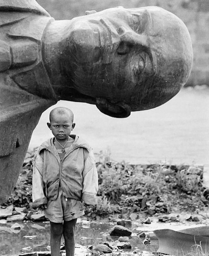 Black and white street photo showing a serious young boy standing near a large fallen statue outdoors.