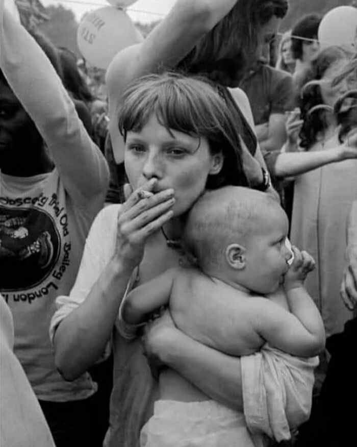 Black and white street photo of a woman smoking while holding a baby amid a crowd, showcasing candid street photography style.