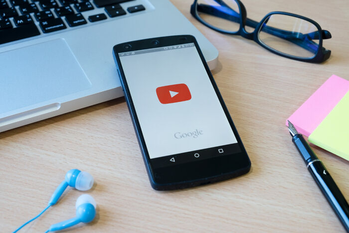 Smartphone with YouTube app open on desk, next to a laptop, earbuds, and colorful sticky notes.