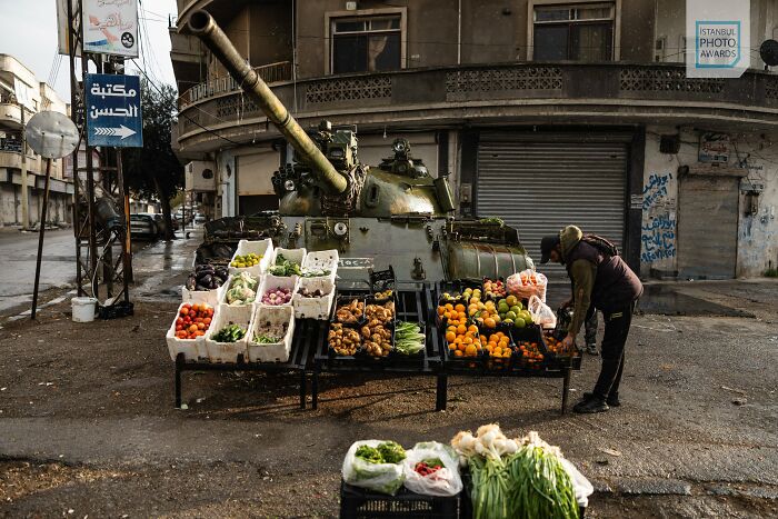 A man sells vegetables from a stand next to a tank in a street, showcasing a scene from the Istanbul Photo Awards 2025.