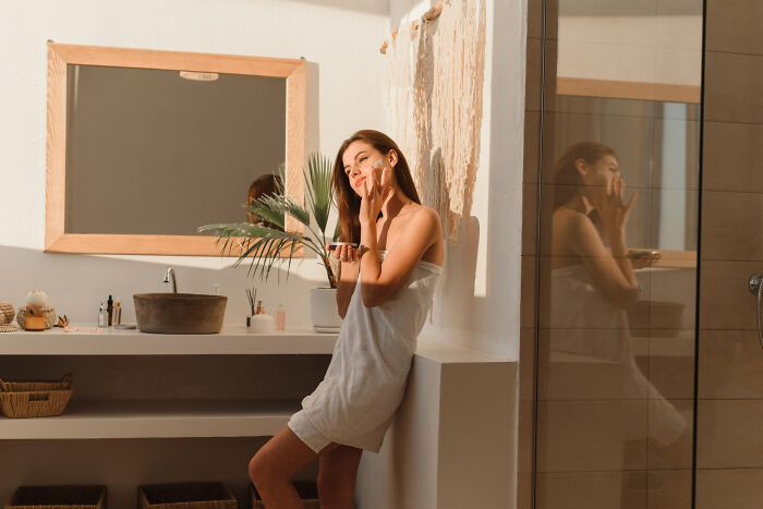 Woman in bathroom wrapped in towel, applying skincare, reflecting on beauty routines.