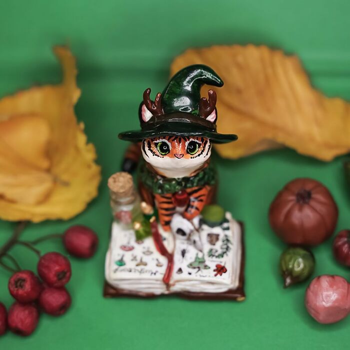 Pocket-sized animal figurine of a cat wizard with a hat, surrounded by colorful leaves and fruits.