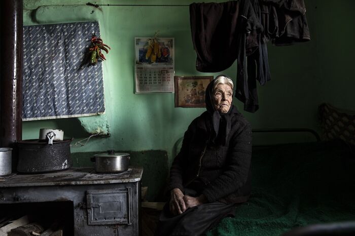 Elderly woman sitting in a dimly lit room, conveying the vulnerability of aging through her expression and surroundings.