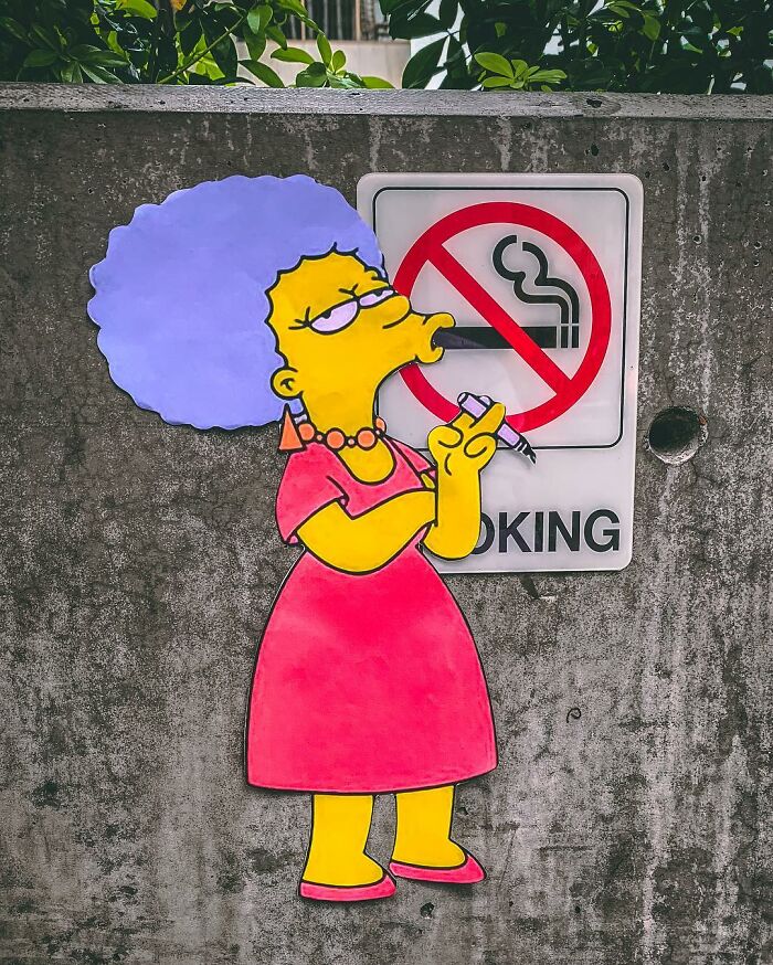Simpsons paper cutout character humorously interacts with a no-smoking sign on a concrete wall.