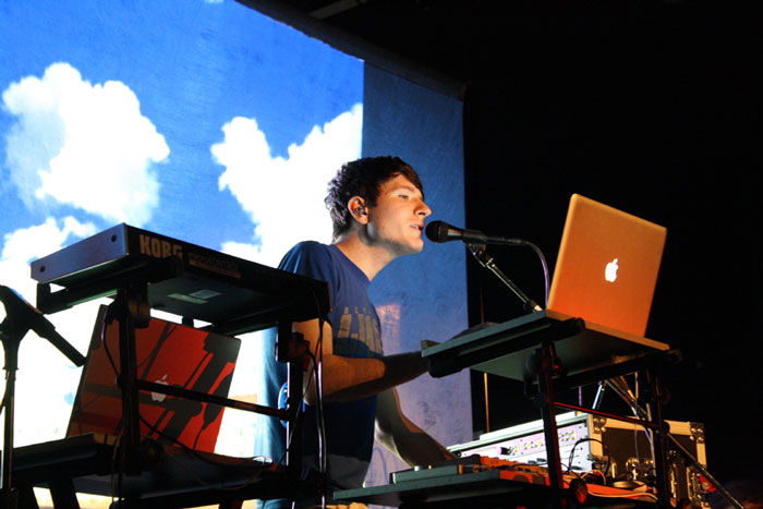 Musician performing live with synthesizers and laptop against a background of clouds.