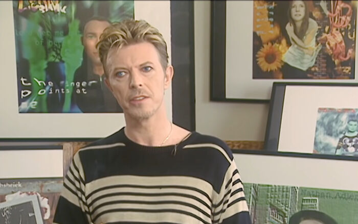 A celebrity in a striped shirt stands in front of framed art, embodying kindness and charm.