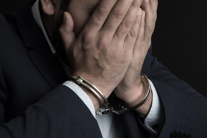 Man in handcuffs covering face with hands against dark background, illustrating intense work drama.