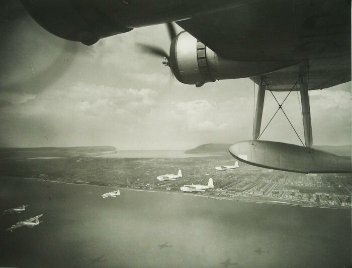 WWII photography captures planes flying over a coastline, viewed from an aircraft wing in black and white.