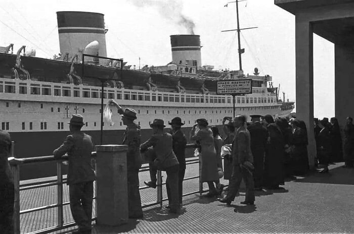 People in early 20th-century attire standing by a pier watching a large ship docked, showing life in Europe 100 years ago.