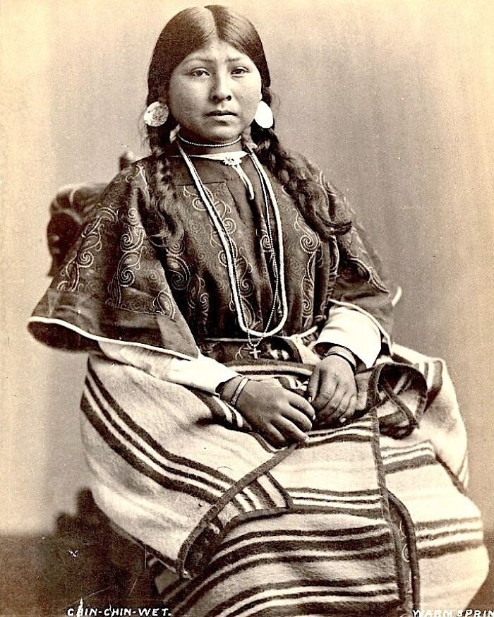 Native American woman from a century ago wearing traditional clothing and jewelry, sitting for a formal portrait.