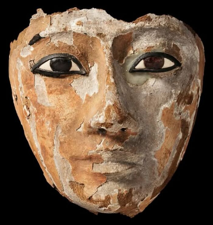 Ancient painted funerary mask with detailed eyes and worn surface found in archaeological excavation site.