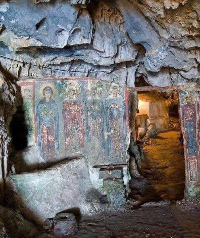 Ancient cave shrine with detailed frescoes of saints inside a historic archaeological site with rock walls and dim lighting.