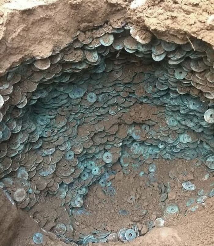 Ancient coin hoard uncovered in archaeological excavation showing thousands of coins stacked in a dirt pit.