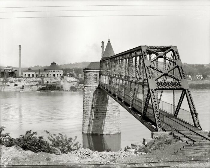 Historic iron bridge crossing a river with industrial buildings in the background, showing life in America 100 years ago
